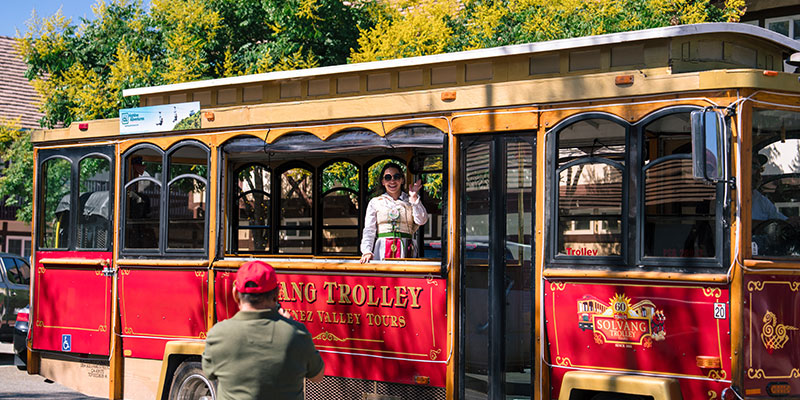 A person in traditional attire waves from a red Solvang Trolley as another in a red cap takes their photo on a sunny day—capturing one of the classic things to do in Solvang, framed by green trees in the background.