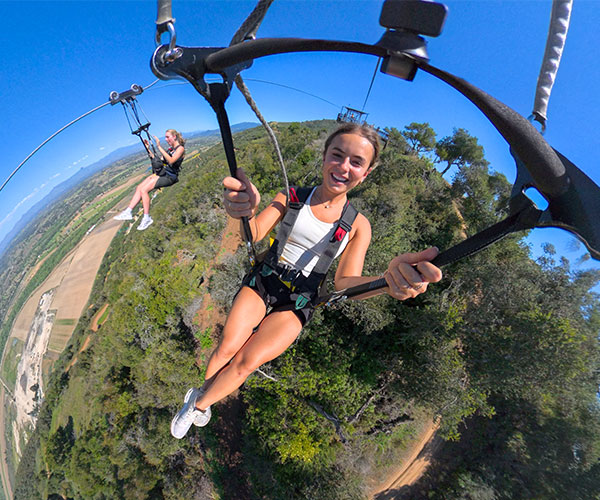 Two women wearing harnesses are ziplining high above a green forest with a scenic landscape in the background. For those seeking adventure, this is one of the thrilling things to do in Solvang on a clear, sunny day. The woman in front smiles excitedly at the camera.