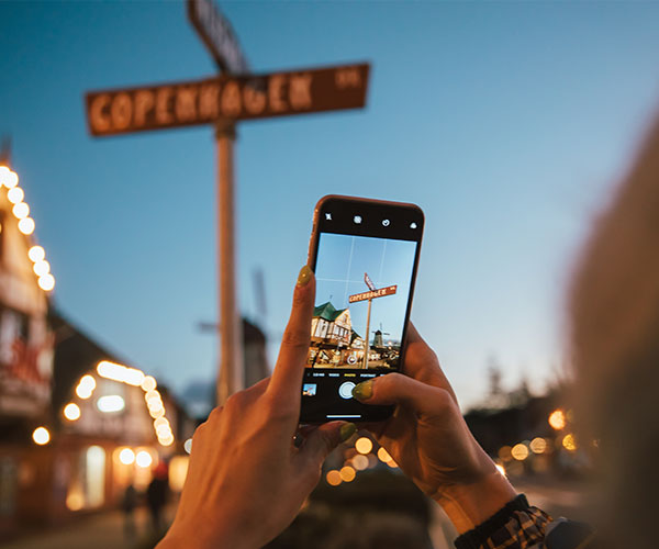 A person holds a smartphone, taking a photo of a street sign that says Copenhagen at dusk—perfect inspiration for Things To Do In Solvang. The background features blurred lights and buildings, creating a cozy evening atmosphere.