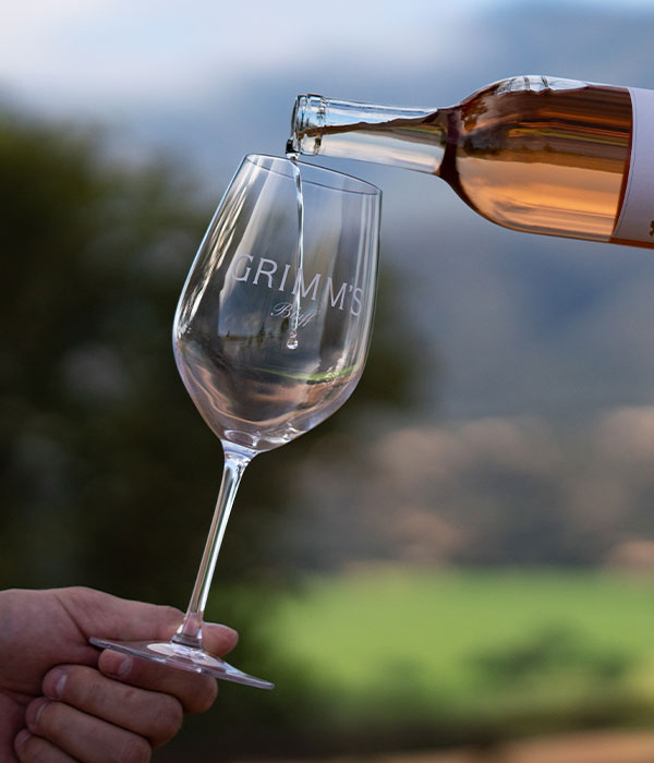 A hand holds a wine glass labeled GRIMMS as rosé is poured from a bottle, with a blurred outdoor landscape in the background—a perfect snapshot of Things To Do In Solvang.