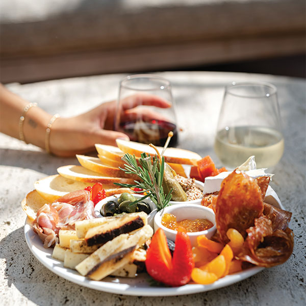 A hand holds a glass of red wine beside a plate of cheeses, meats, fruits, crackers, olives, and dip. Another glass of white wine sits in sunlight—perfect for savoring with Special Offers at The Landsby.