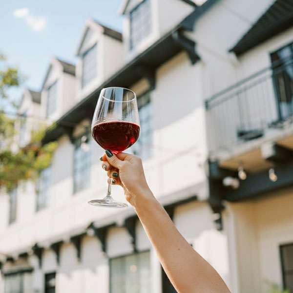 A hand with dark nail polish holds up a glass of red wine outdoors in front of a white building with multiple windows—perhaps while enjoying Special Offers at The Landsby—under a clear blue sky.