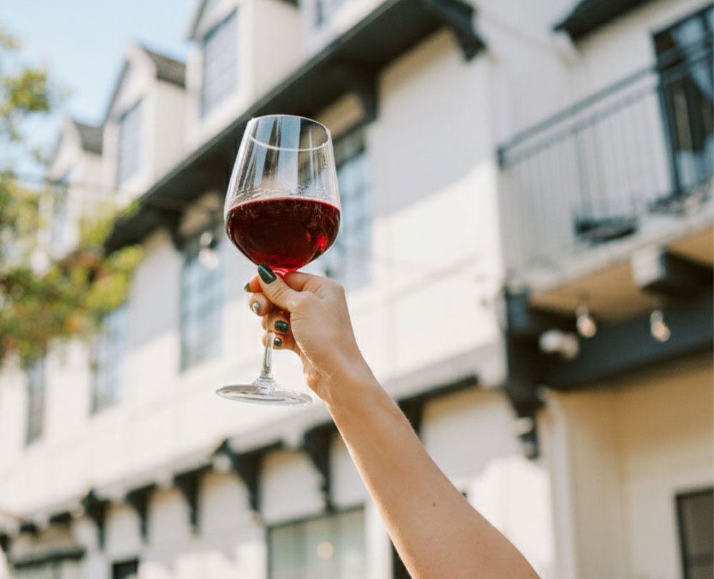 A hand with painted nails holds up a glass of red wine outdoors at a boutique hotel in Solvang, with a blurred background of a white building and a sunny sky.