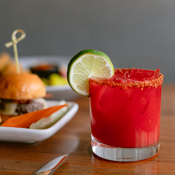 A vibrant red cocktail in a glass rimmed with chili powder, garnished with a lime slice, sits on a wooden table next to sliders and pickles—an inviting scene for those seeking Top Solvang Dining experiences.