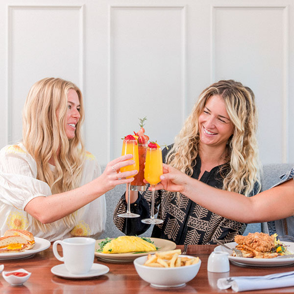 Three women smiling and clinking glasses filled with colorful drinks over a table set with breakfast foods—omelets, fries, pancakes, and fried chicken—enjoy the morning at one of the top Solvang dining spots.