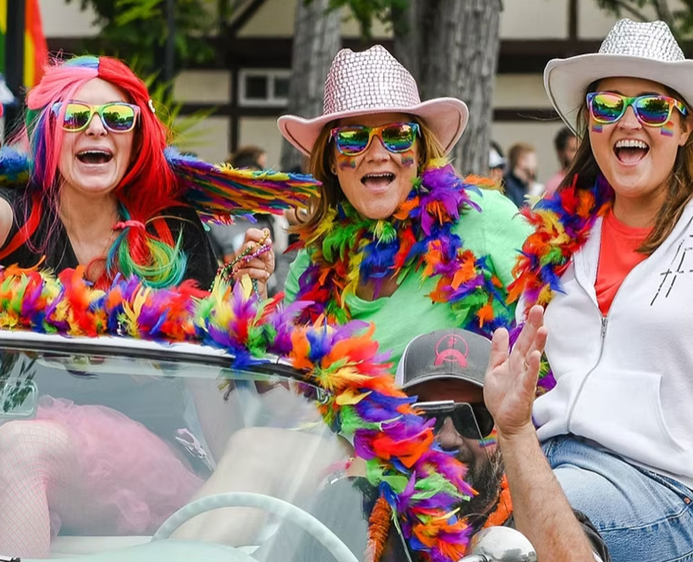 Three people in colorful feather boas, wigs, and rainbow sunglasses smile and cheer in a lively parade convertible—just one of the fabulous things to do in Solvang. A man in a cap waves joyfully in front of them, adding to the festive scene.