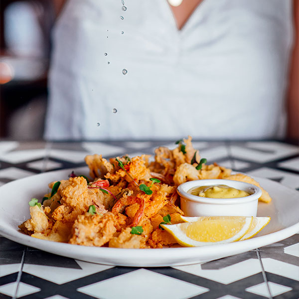 A plate of fried calamari garnished with herbs, served with lemon wedges and a small dish of dipping sauce, sits on a patterned table—just one example of Top Solvang Dining enjoyed by a guest in a white shirt.