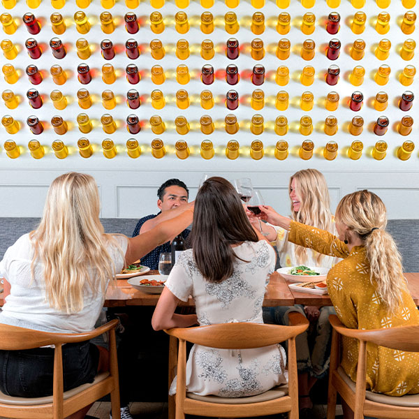 Five people sit around a dining table toasting with drinks and enjoying plates of food, surrounded by rows of colorful glasses or jars—a perfect evening at one of the top Solvang dining spots.