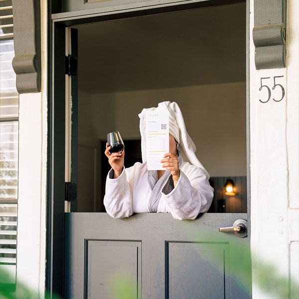 A person in a bathrobe stands behind a half-open door marked 55, sipping red wine and covering their face with a book—perhaps unwinding after exploring Special Offers at The Landsby.