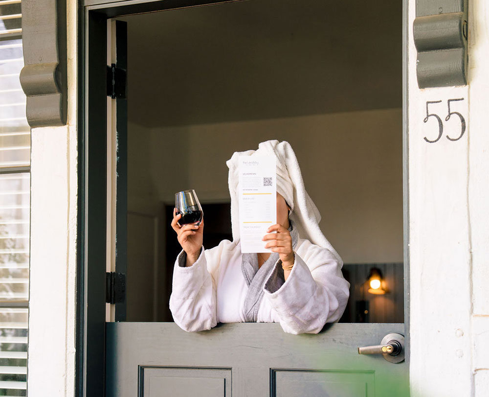 A person in a white bathrobe and towel holds up a wine glass and a piece of paper featuring Special Offers at The Landsby, standing at a half-open door with the number 55 visible. Their face is hidden behind the paper.