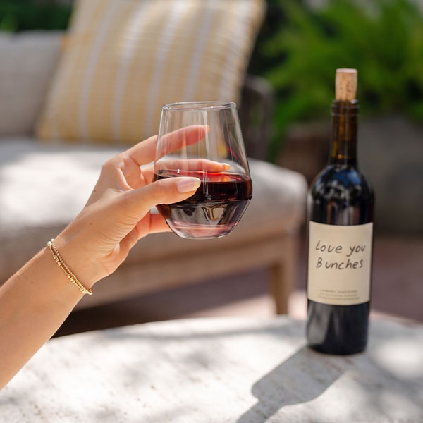 A hand holding a glass of red wine near a table, with a bottle labeled love you Bunches in the background. The outdoor patio setting, sunlight, and greenery evoke the relaxed charm of Top Solvang Dining experiences.