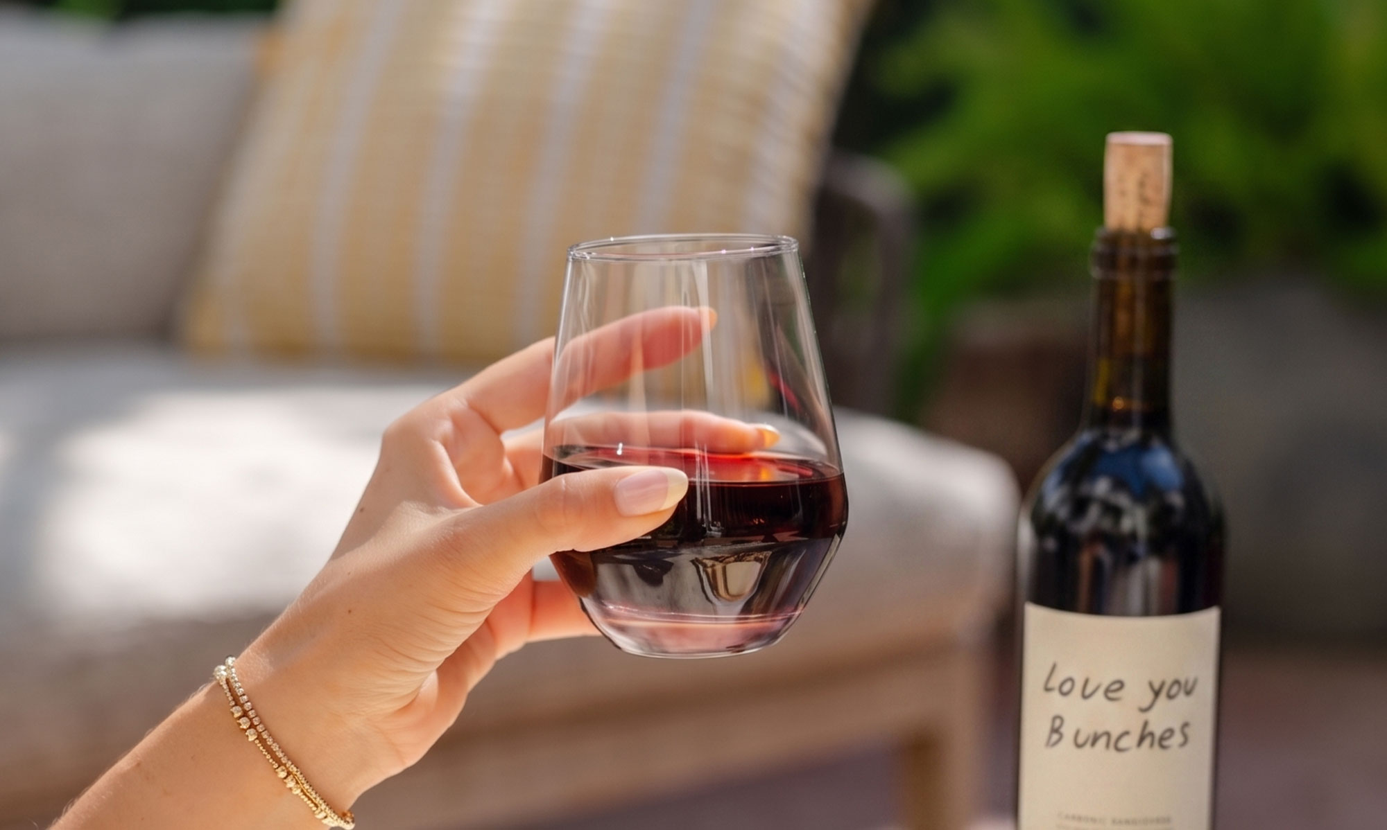 A hand holding a glass of red wine outdoors with a bottle labeled Love you Bunches nearby; Hotel Happenings at The Landsby set the scene with a cushioned chair and greenery softly blurred in the background.